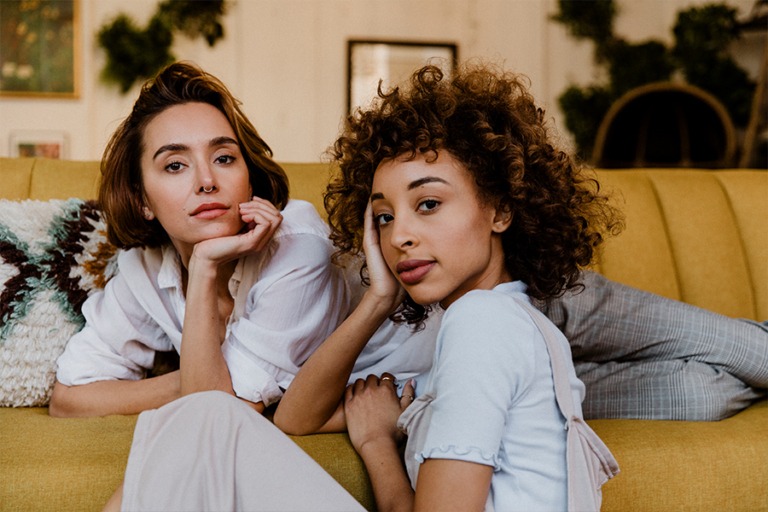 Woman laying on yellow couch in modern Millennial apartment, facing the camera with friend sitting on floor also facing camera