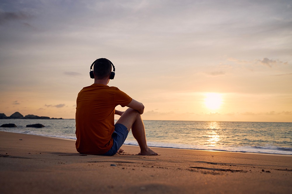 Man listening to music on cans style headphones while sitting on the beach during an end of year reflection