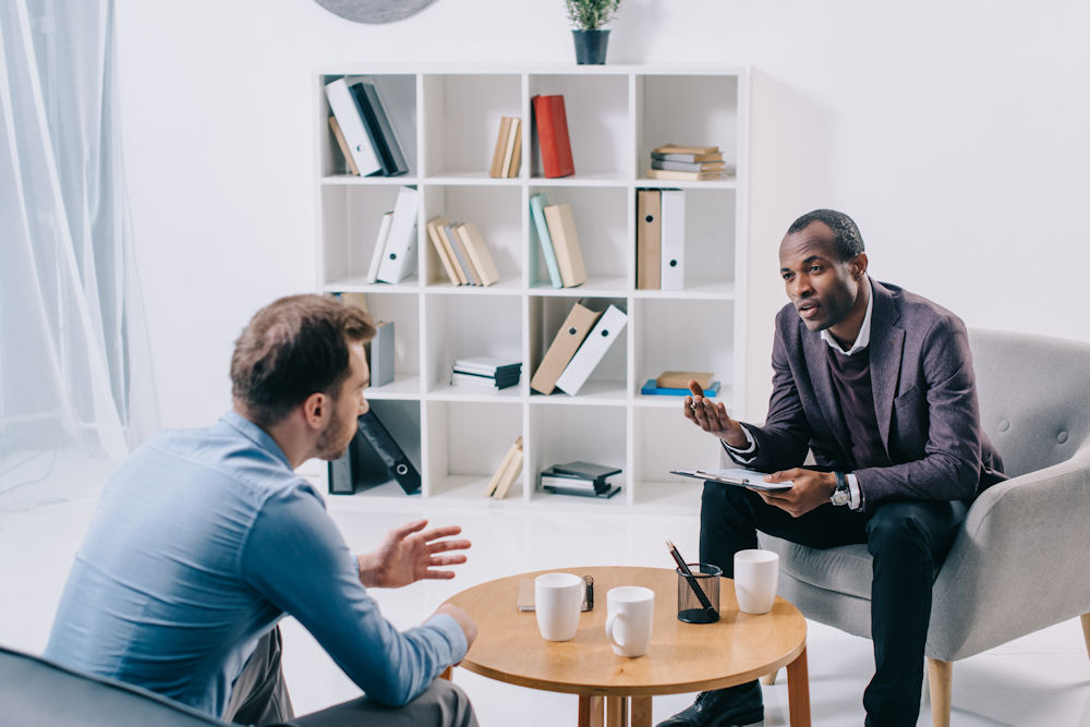 Therapist in all-white office office explaining triggers in recovery to male patient asking questions