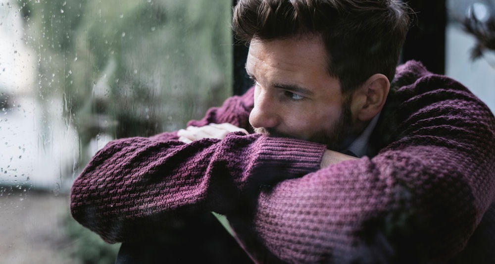 Man folded arms and while looking out a rainy window during depression relapse