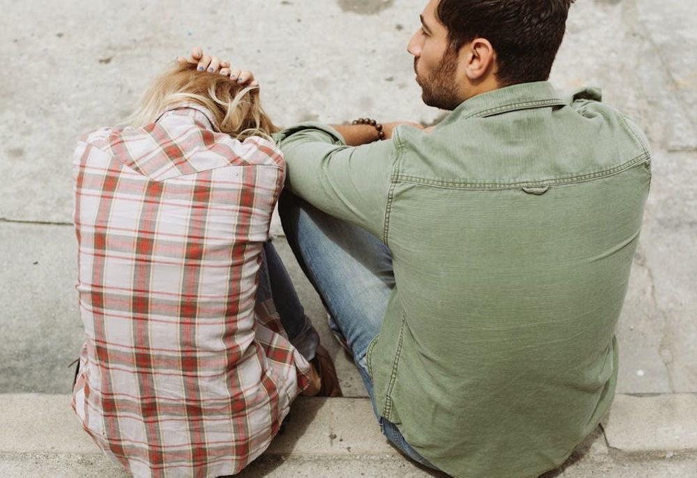 Woman holds head in hands as man watches on, both sitting on stone steps